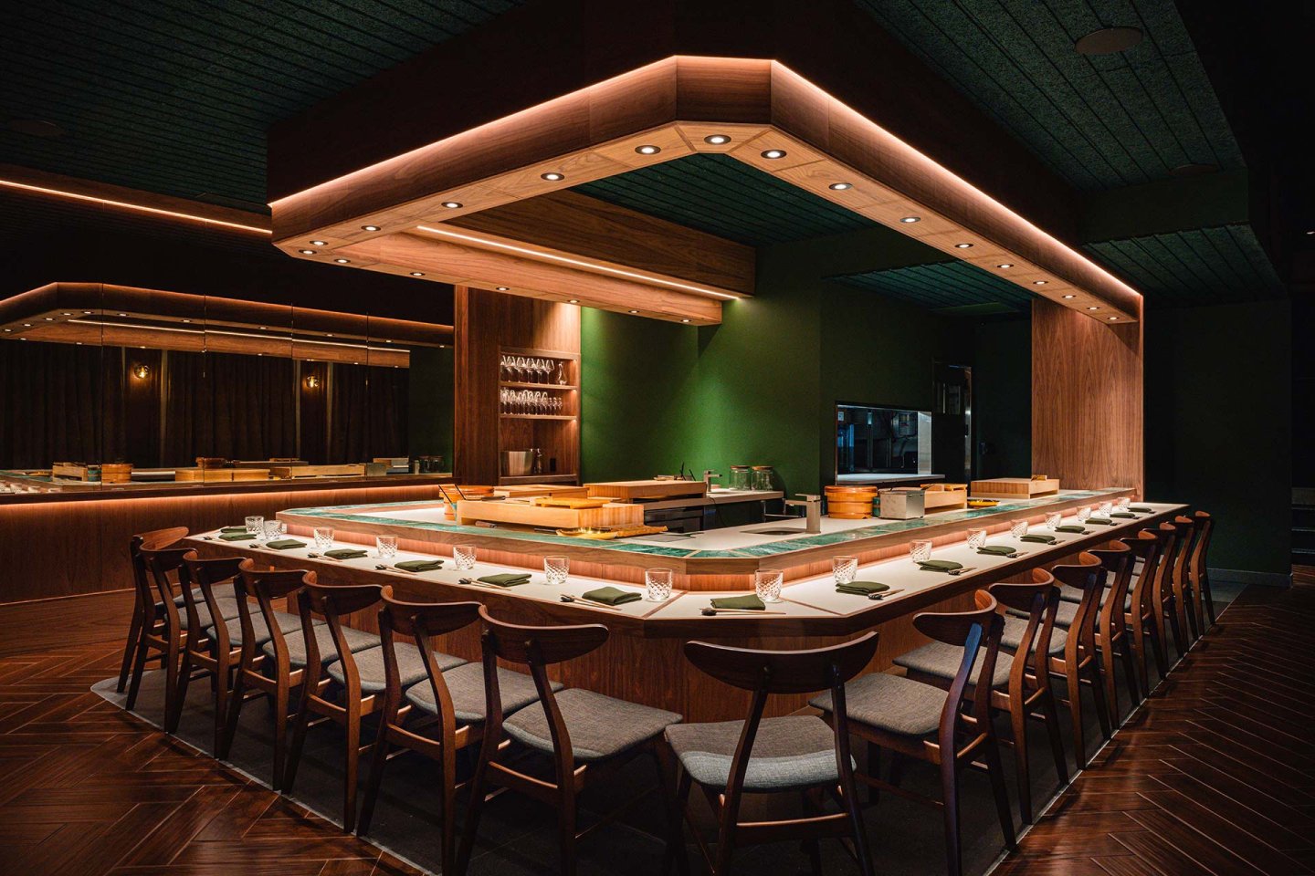 A modern sushi bar with a U-shaped wooden counter surrounded by neatly arranged wooden chairs, set with plates and glassware. Warm lighting highlights the sleek, minimalist decor and green walls.