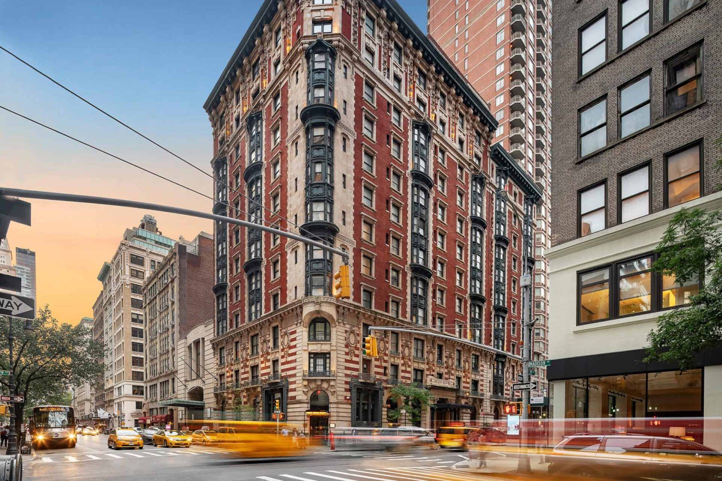 A busy New York City street corner at sunset with a historic red-brick apartment building, yellow taxis, and blurred motion from traffic and people.