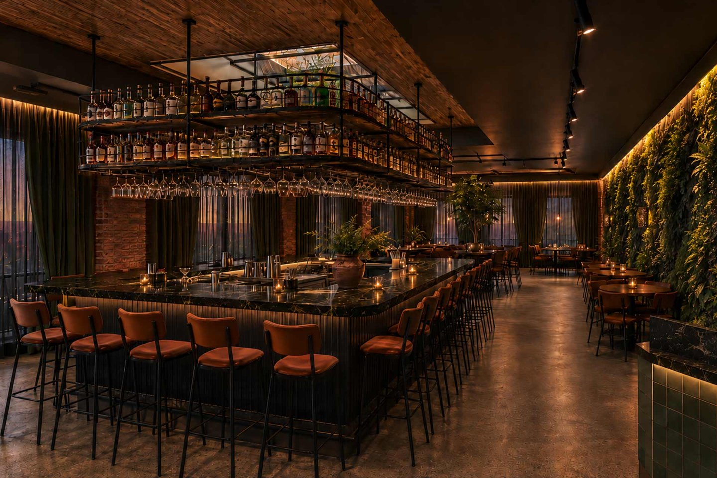 Chic, modern bar with a large black counter, brown leather stools, hanging glassware, shelves of liquor, dim lighting, and a lush green plant wall along one side. Tables and chairs fill the rest of the stylish space.