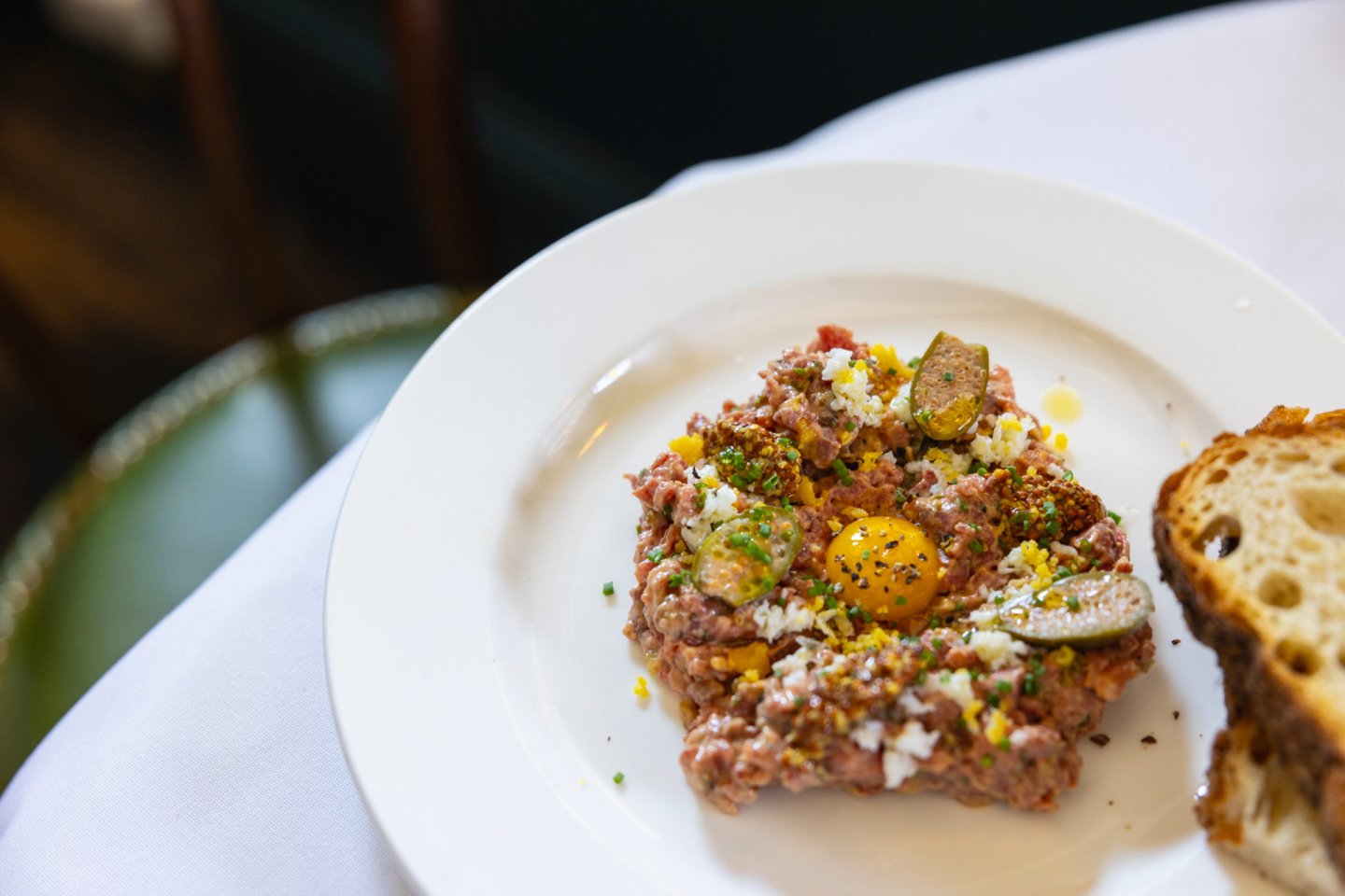 A plate of beef tartare topped with a raw egg yolk, chopped herbs, and capers, served with slices of rustic bread on a white tablecloth.
