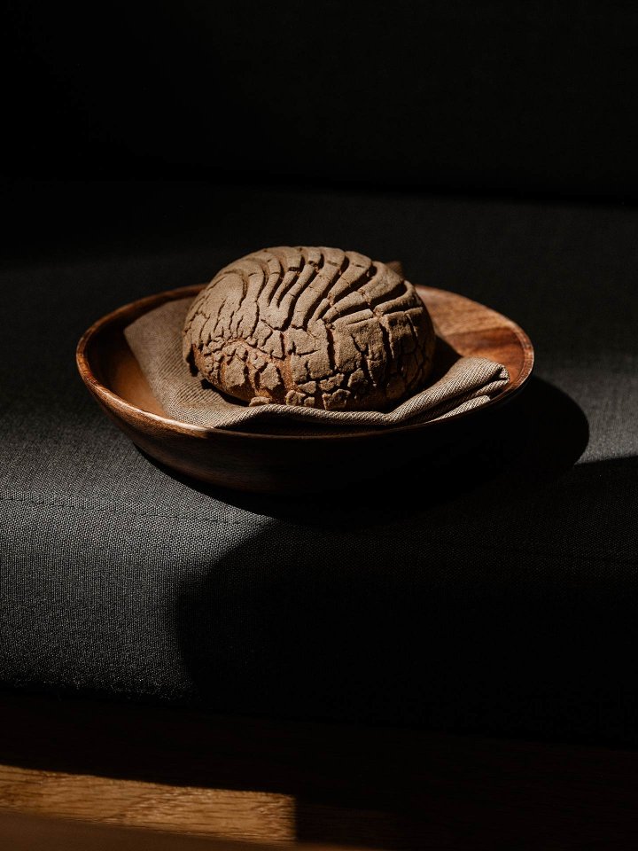 A round loaf of dark bread with a textured crust sits on a cloth napkin in a wooden bowl, placed on a dark surface with dramatic lighting and deep shadows.