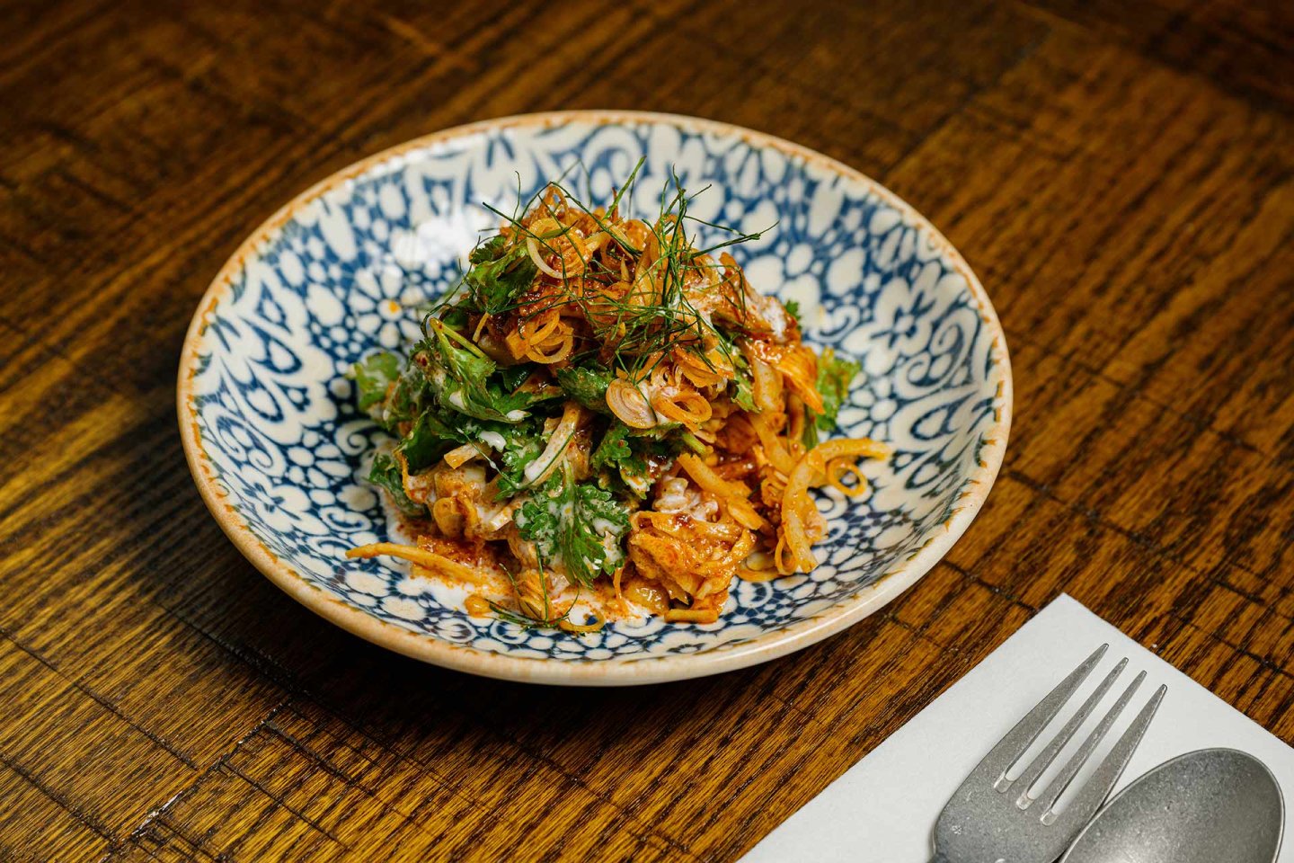 A blue and white patterned bowl filled with a colorful salad of herbs, sliced vegetables, and a red spicy sauce sits on a wooden table next to a fork and spoon on a napkin.