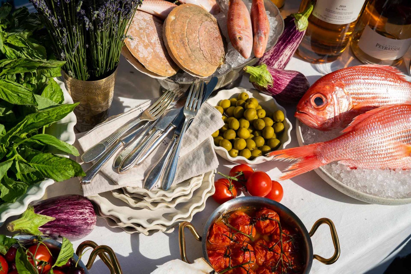 A table with fresh red fish, clams, eggplants, basil, green olives, tomatoes, cutlery, a dish of roasted tomatoes, bottles of wine, and a vase of lavender on a white tablecloth in bright sunlight.