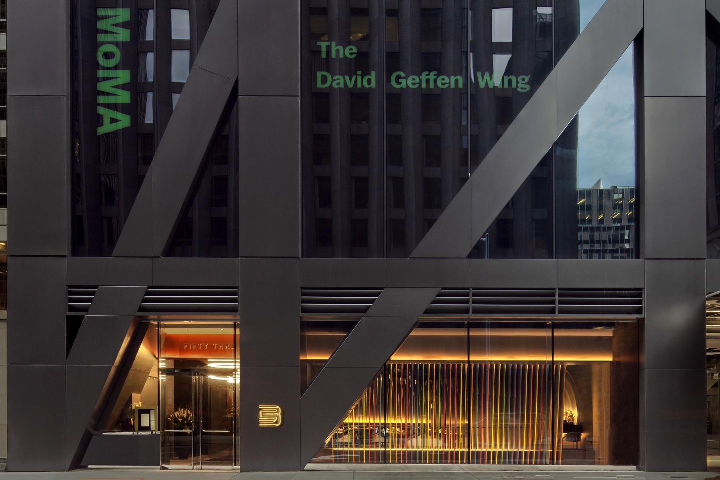 The exterior of the Museum of Modern Art (MoMA) David Geffen Wing, featuring modern architecture with large glass panels, bold black framing, and illuminated interior visible through the entrance.