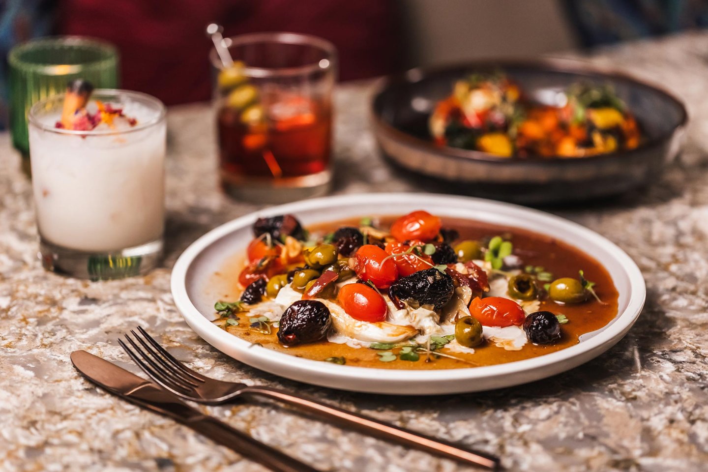 A plate of roasted vegetables, cherry tomatoes, green olives, and cheese sits on a marble table, with a fork and knife beside it and two cocktails and another dish in the background.