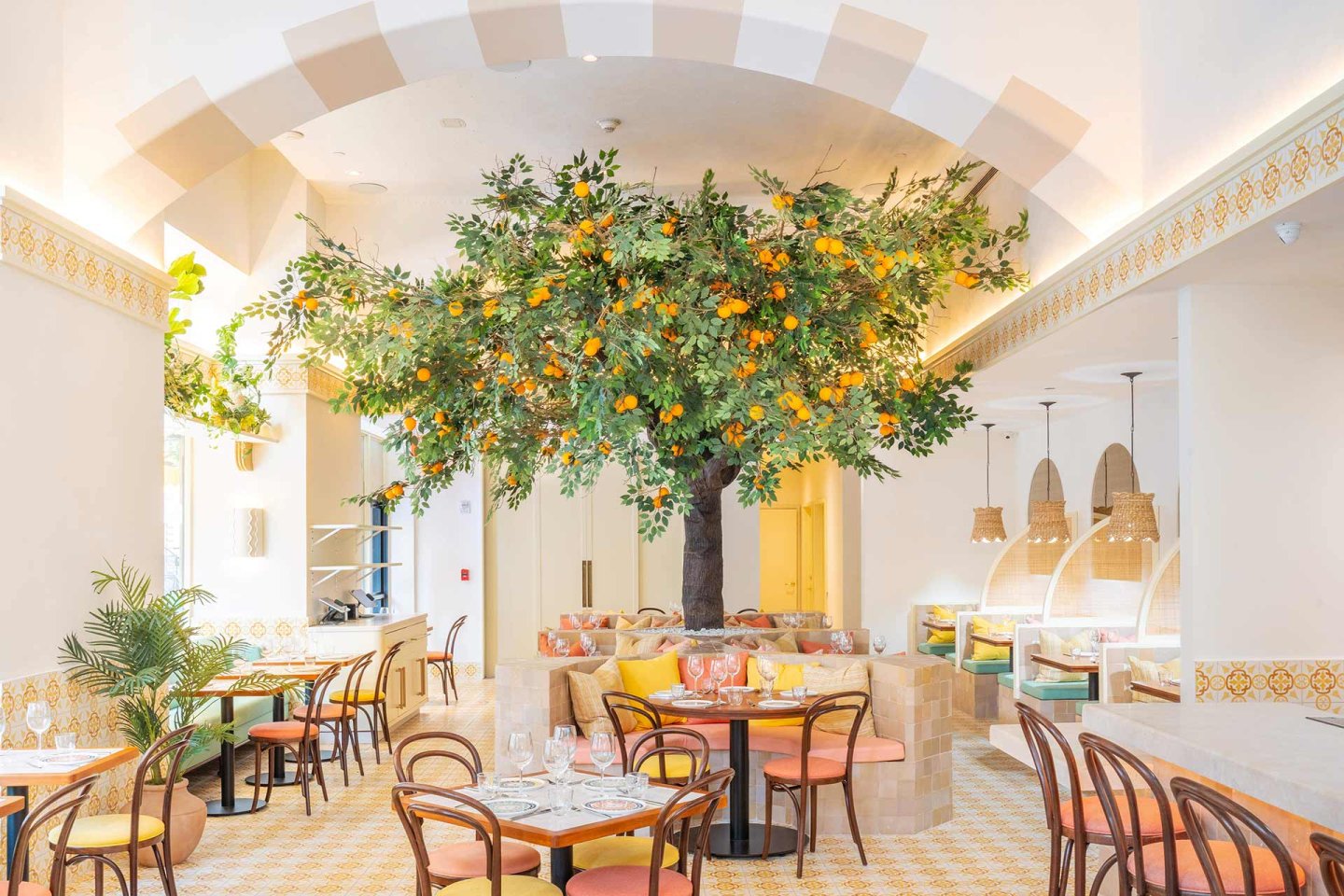 A bright, airy restaurant interior features a large artificial orange tree as its centerpiece, surrounded by tables and chairs, woven basket light fixtures, and pastel-colored decor with Mediterranean-inspired patterns.