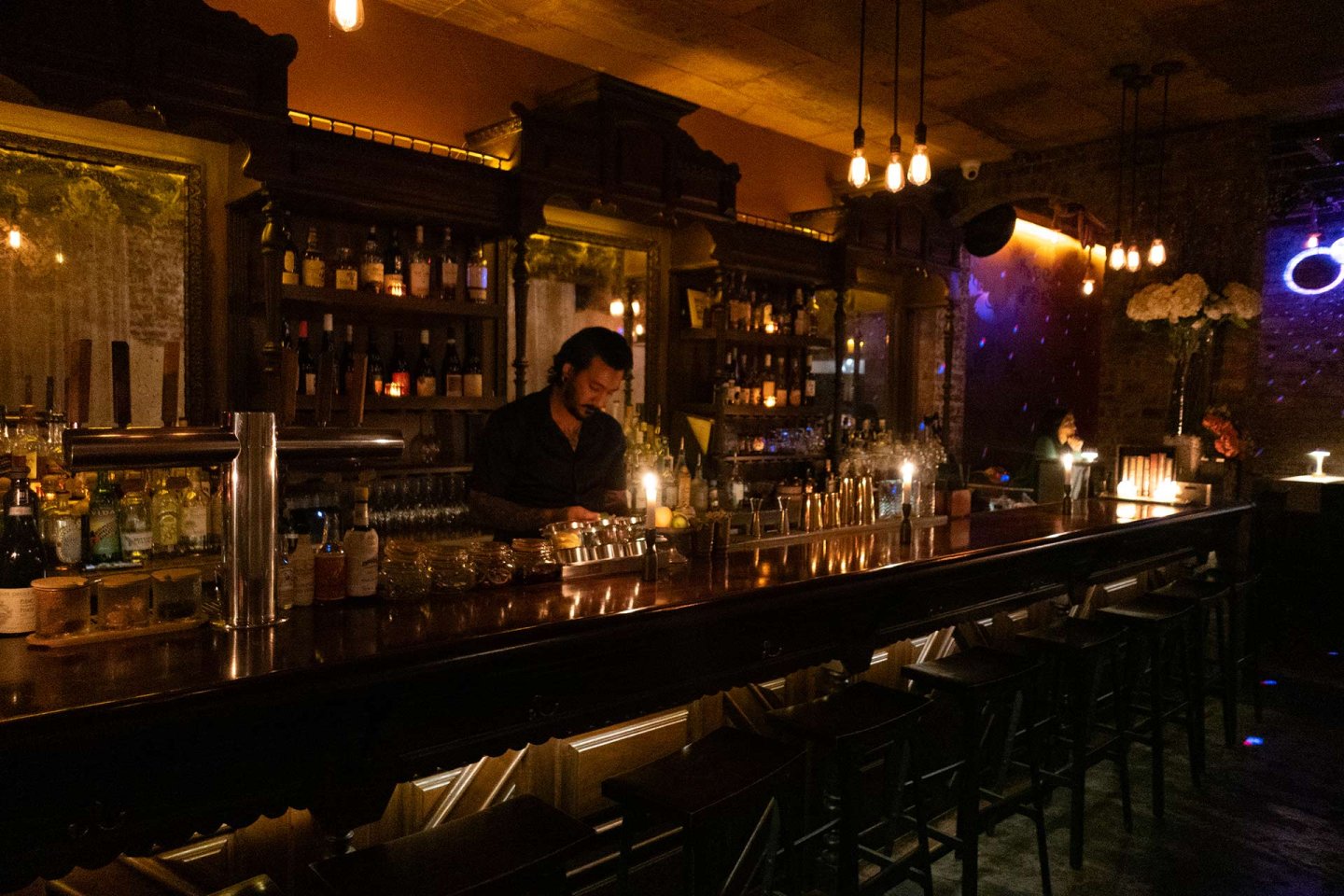A bartender prepares drinks behind a dimly lit bar with shelves of bottles and glasses, surrounded by dark wood, warm lighting, and empty bar stools in an intimate, cozy atmosphere.