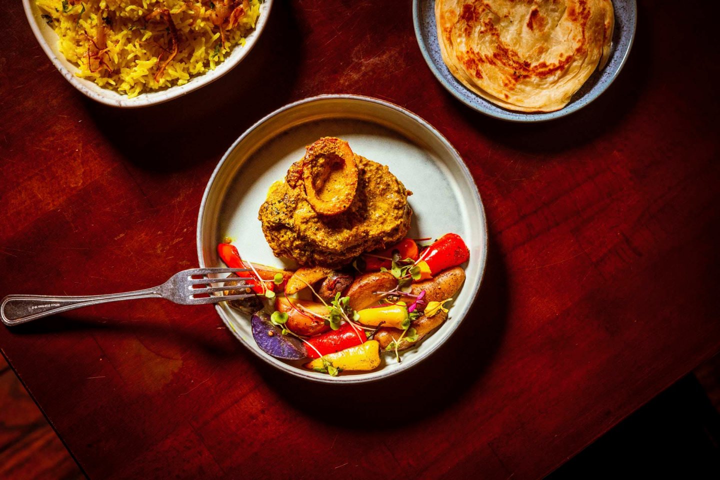 A plate with a portion of meat, garnished with herbs, and assorted colorful vegetables. A fork is placed on the plate. In the background, there are side dishes of rice and bread on a wooden table.