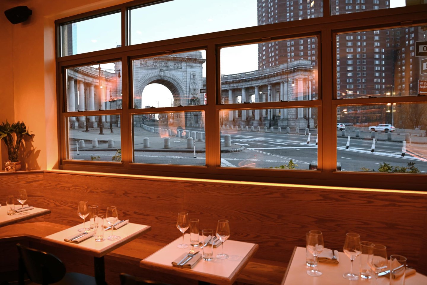 Restaurant tables set with glassware and napkins by a window, overlooking a city street and a large stone arch structure outside at dusk.