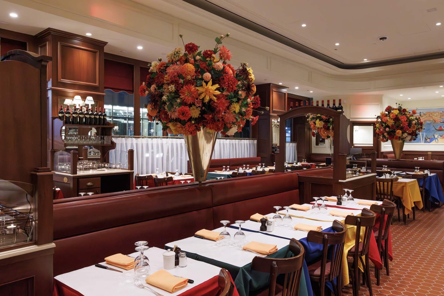 A spacious, elegant restaurant with wooden booths, colorful tablecloths, large floral centerpieces, set wine glasses, and a warm, inviting ambiance. Shelves with bottles and framed art decorate the background.