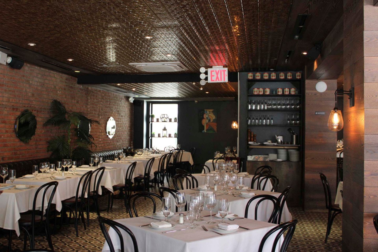 A dimly lit restaurant with neatly set tables covered in white linens. Black chairs surround the tables, and the décor features exposed brick walls, a patterned floor, and a tin ceiling. A small bar area is visible in the background.