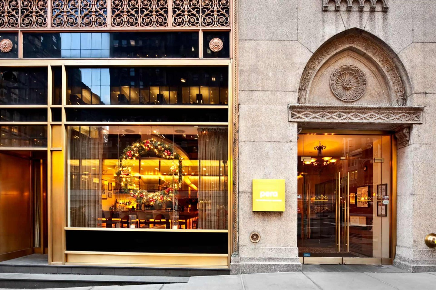 The exterior of a restaurant with large windows, a holiday wreath inside, ornate metalwork above, and a yellow sign near the entrance reading “pera.”.