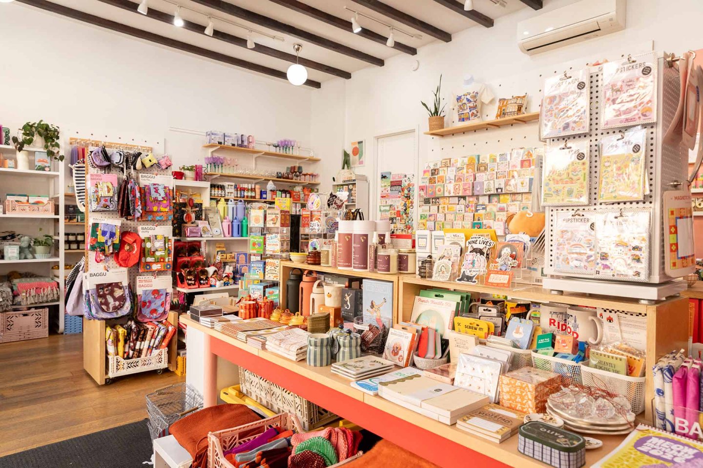 Bright stationery shop interior with colorful notebooks, pens, greeting cards, art supplies, and gift items displayed on shelves and tables, with wooden floors and white walls.