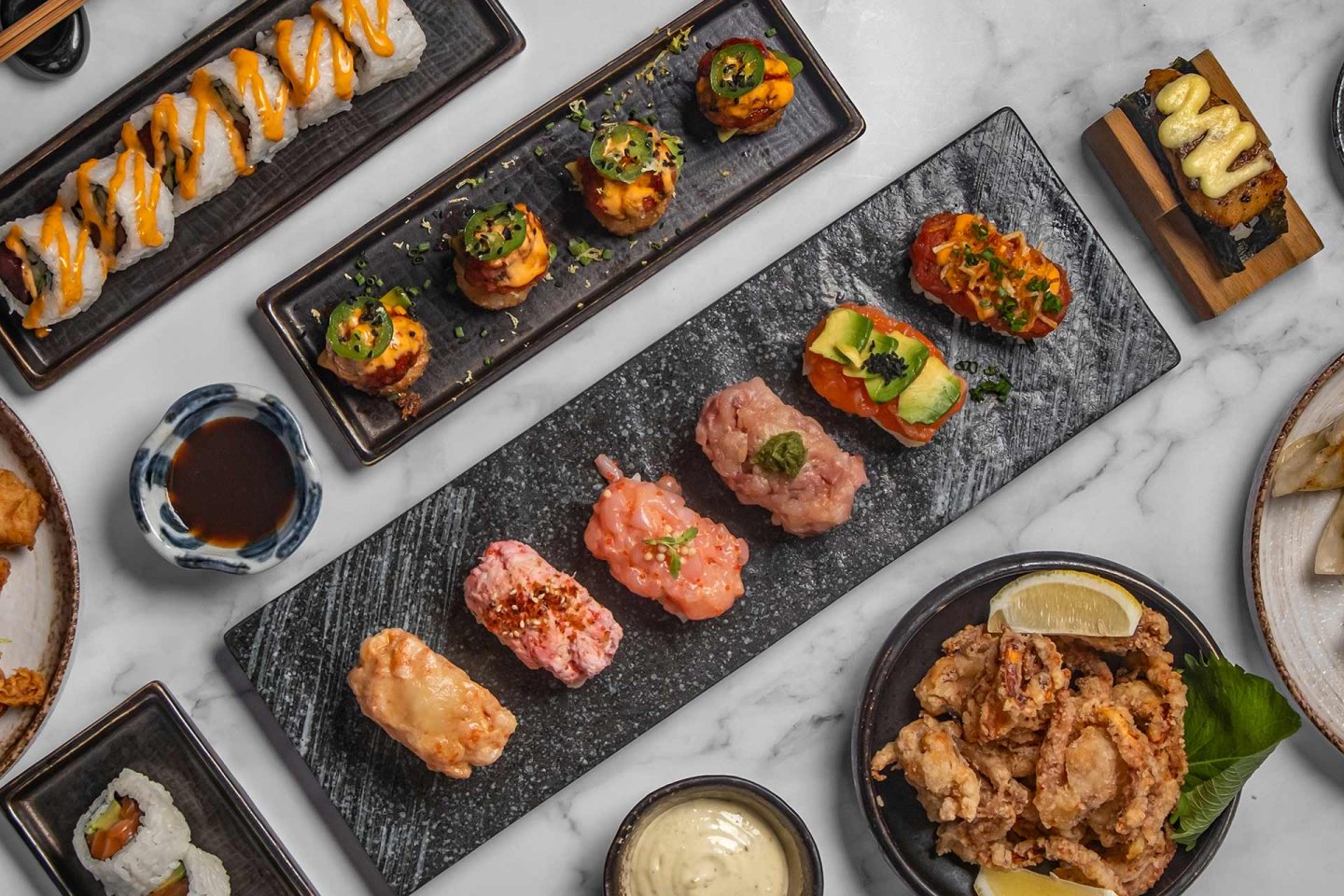 Assorted sushi, sashimi, and nigiri arranged on black trays, with fried appetizers, dipping sauces, and garnishes on a marble table.