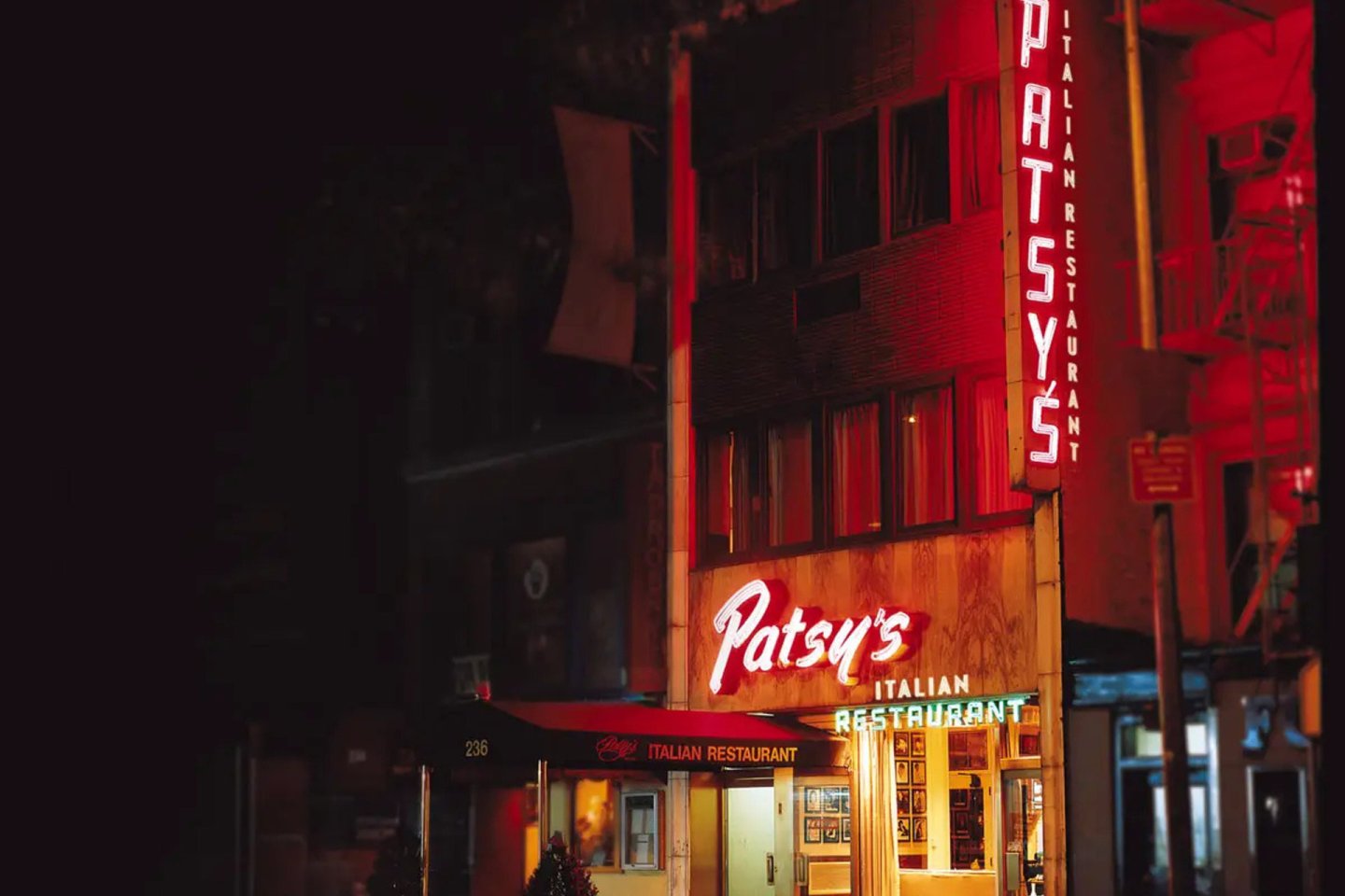 A warmly lit street view of Patsy's Italian Restaurant at night, featuring illuminated red and white signs on a dark building facade.