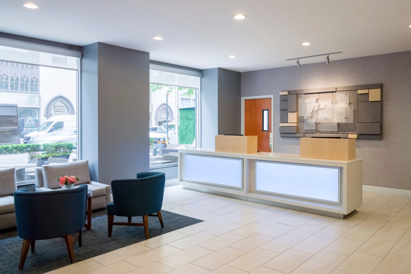 A modern hotel lobby with a reception desk. The space features bright lighting, contemporary furniture, and large windows. Two upholstered chairs and a small table with flowers are on the left, while the reception area is on the right.