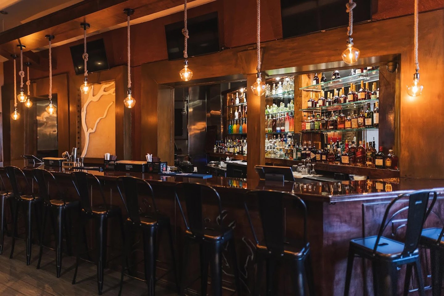 A stylish, empty bar with metal stools, pendant bulbs hanging above, and shelves stocked with various bottles of alcohol behind the wooden counter. The atmosphere is warm and inviting.