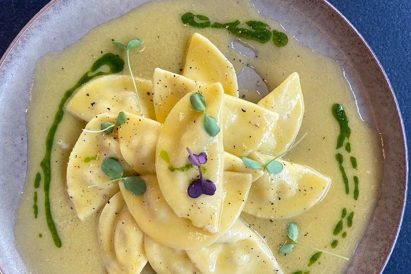 A plate of ravioli arranged in a circular pattern, served with a creamy yellow sauce, garnished with microgreens and a drizzle of green herb oil.