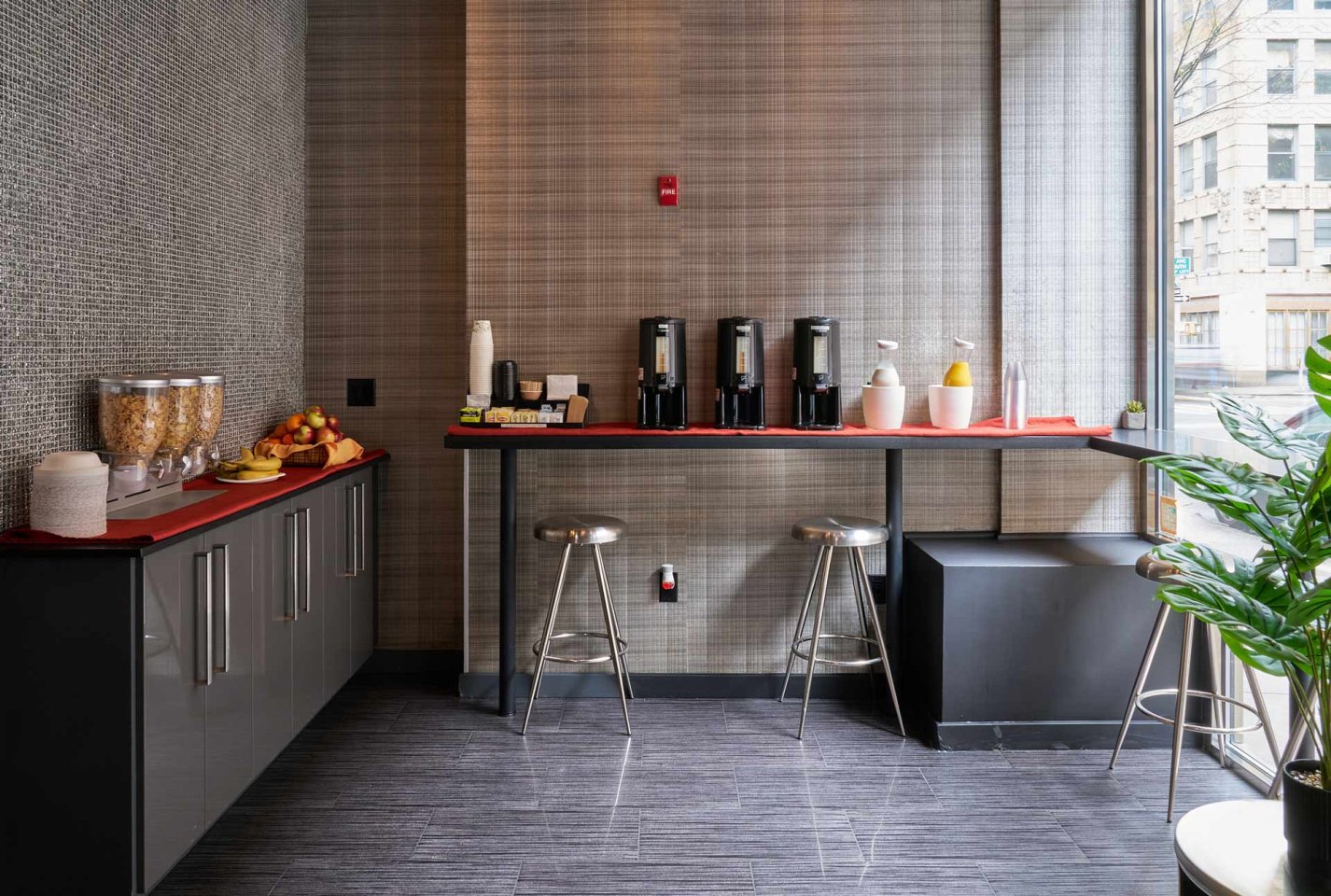 A modern breakfast area with cereal dispensers, fruit, cups, coffee urns, condiments, and two metal stools at a counter by a window overlooking city buildings.