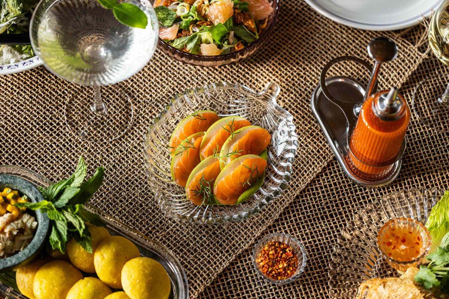 A glass dish with four salmon sashimi pieces garnished with herbs is on a woven tablecloth, surrounded by various dishes, a drink, condiments, and fresh greens.