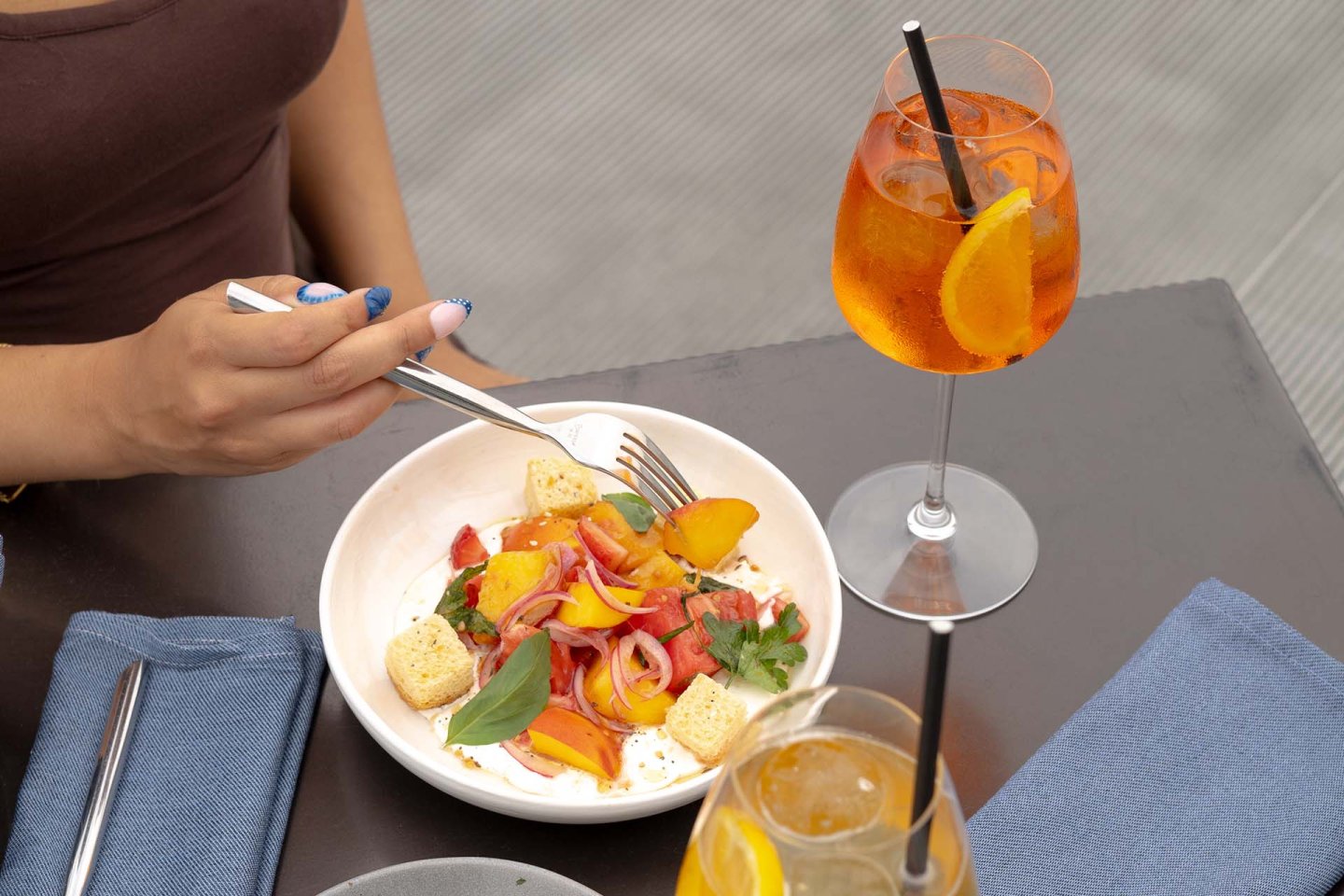 A person prepares to eat a fresh salad with tomatoes, peaches, onions, croutons, and herbs, next to two orange cocktails on a table with blue napkins.