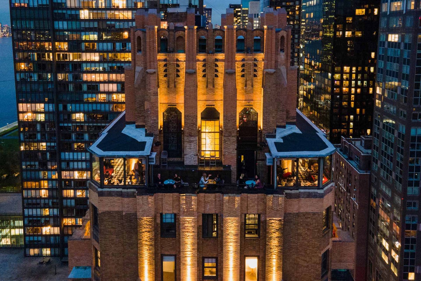 Aerial view of a tall, illuminated brick building with gothic architectural details, surrounded by modern glass skyscrapers at dusk. People are visible dining on terraces. City lights glow in the background.