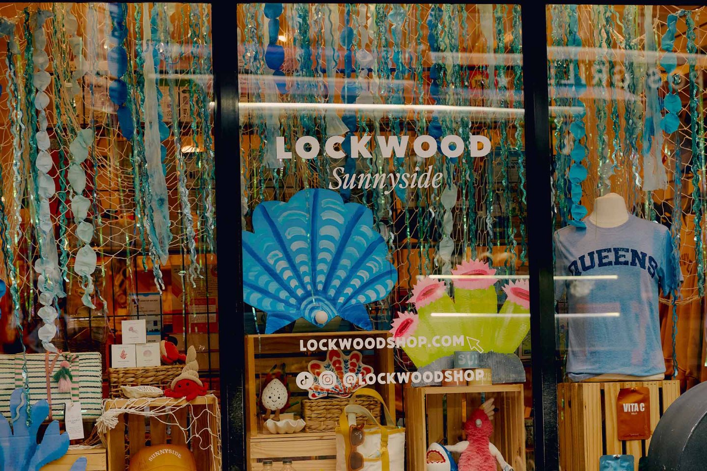 A colorful shop window display at Lockwood Sunnyside features hanging blue and turquoise decorations, a large shell cutout, gift items, books, and a mannequin wearing a light blue "QUEENS" t-shirt.