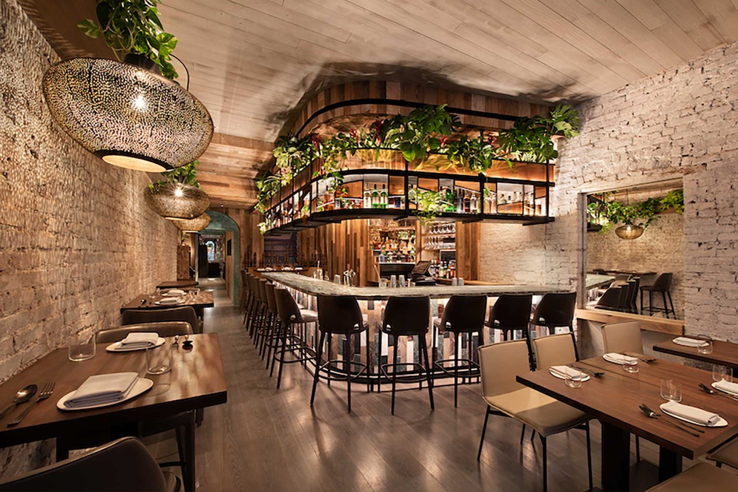 A stylish restaurant interior with exposed brick walls, wooden tables, cushioned chairs, and a curved bar. Hanging plants and ornate light fixtures add warmth and greenery to the elegant, modern atmosphere.