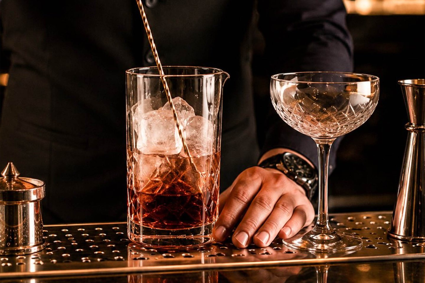 A bartender stirs a cocktail with ice in a mixing glass next to an empty coupe glass on a bar counter, with cocktail tools visible nearby.