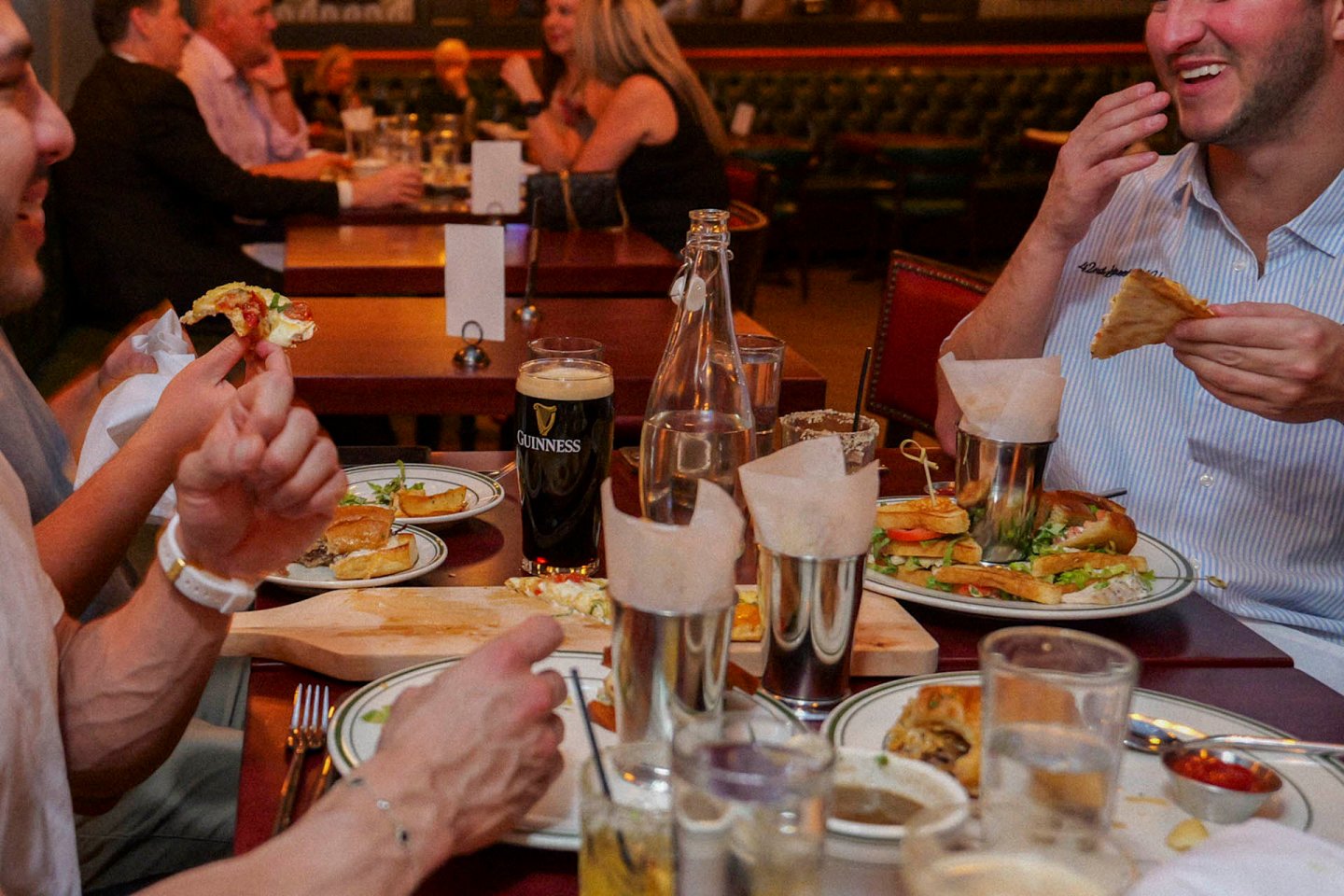 People are sitting at a restaurant table, enjoying a meal and drinks. Plates of food and glasses, including a pint of dark beer, are visible. One person holds a slice of pizza, and another is laughing while holding a sandwich. The atmosphere is lively.