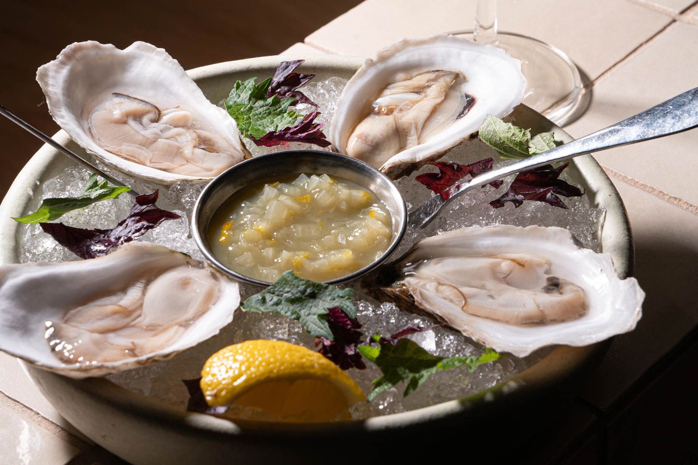 A plate of four raw oysters on the half shell sits on crushed ice with leafy garnishes, a lemon wedge, and a small dish of sauce in the center. A spoon rests on the edge of the plate.
