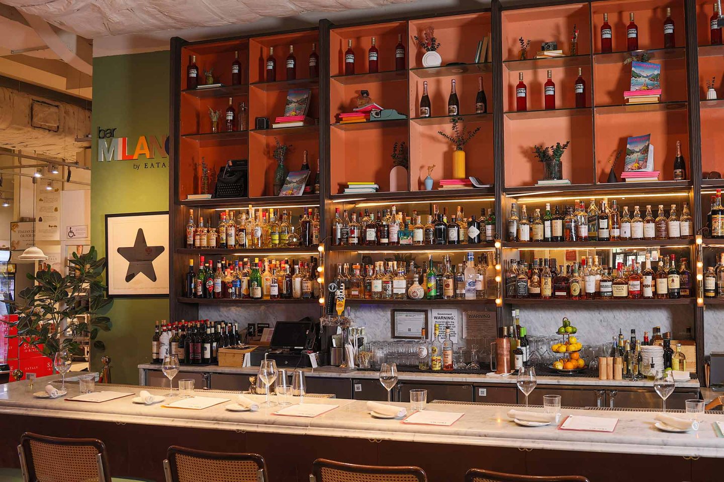 A stylish bar with shelves filled with bottles of liquor and wine, glasses hanging above, a marble counter set with plates and wine glasses, and chairs lined up in front. Warm lighting creates a welcoming atmosphere.