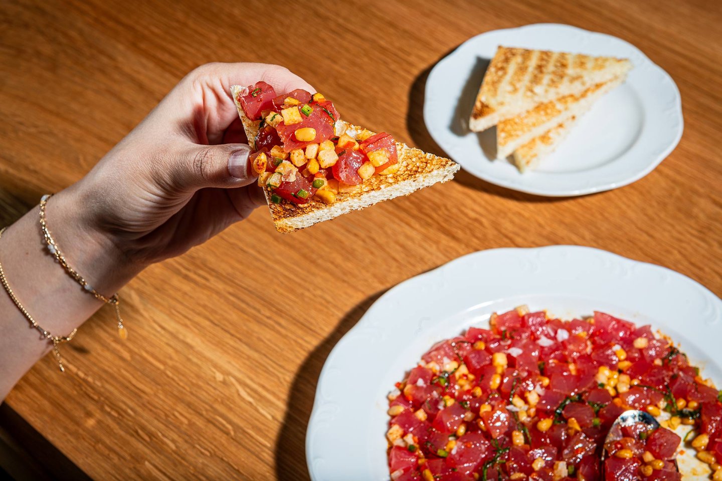 A hand holds a piece of toast topped with diced raw tuna and vegetables. A plate with more tuna mixture and another plate with plain toast triangles sit on a wooden table.