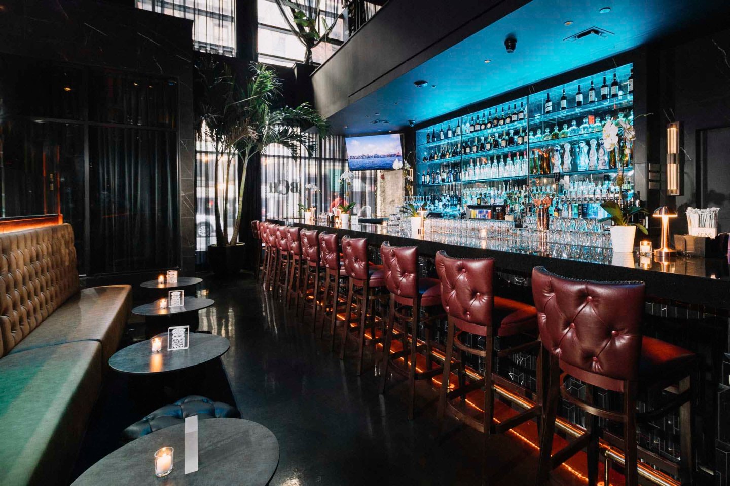 A dimly lit, modern bar with a long counter lined with red upholstered stools. Shelves behind the counter display bottles and glassware, illuminated by blue lighting. Small candles light the tables opposite the bar, creating an intimate atmosphere.