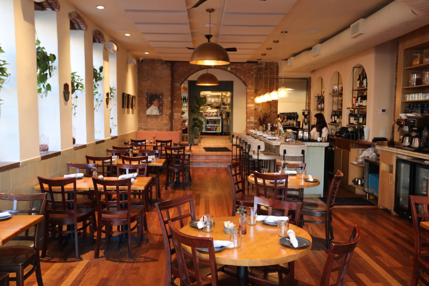 A cozy restaurant interior with wooden floors, round tables set for dining, and chairs arranged neatly. Large windows line one wall, and warm pendant lights hang from the ceiling, creating an inviting atmosphere.