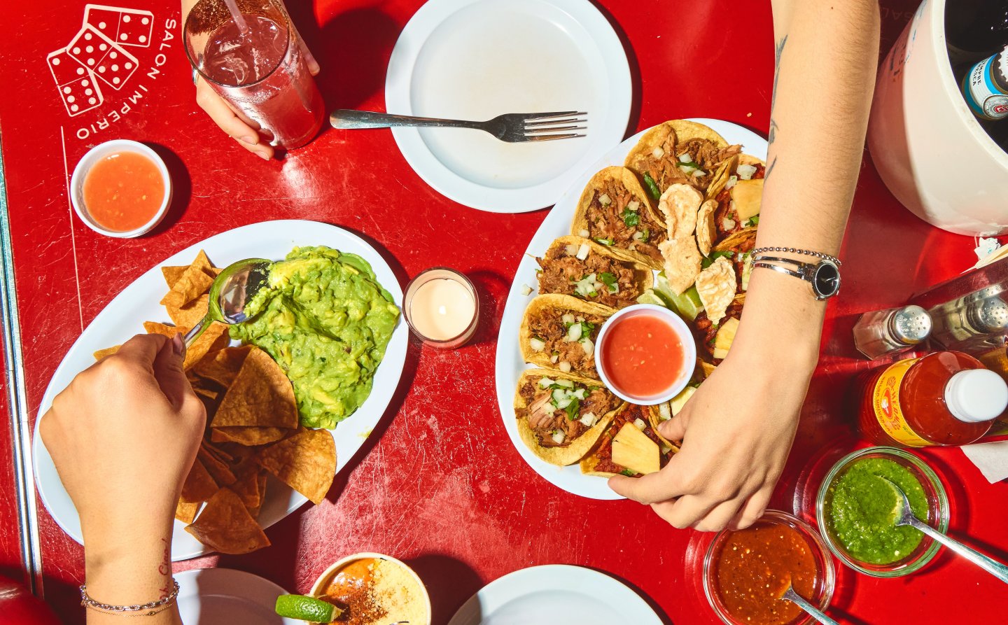 An overhead view of a bright red table filled with tacos, guacamole and chips, salsa, and cocktails. Guests reach across the table to share food, creating a lively, communal dining scene at a bustling taqueria.