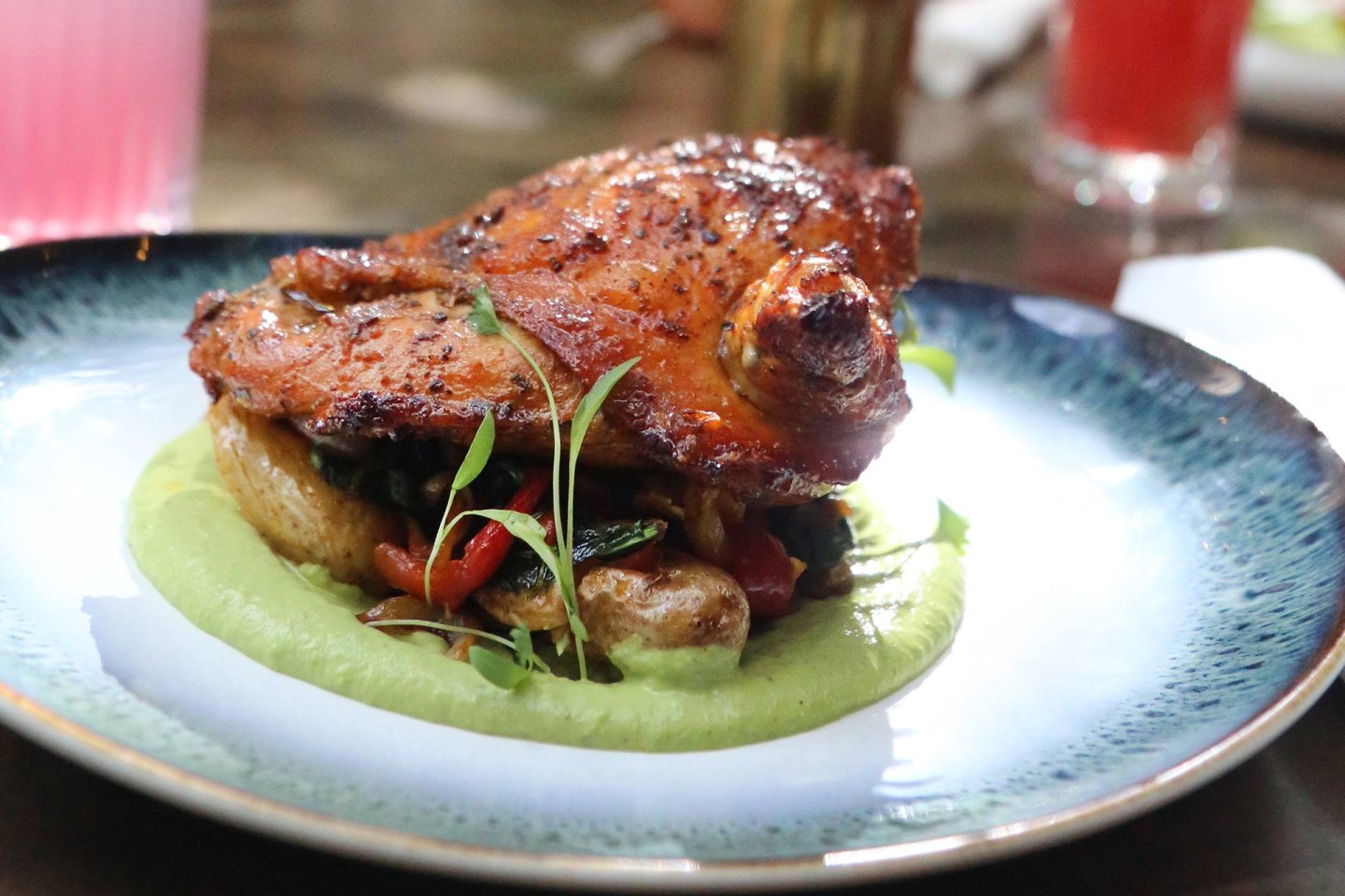 A roasted chicken thigh served on a bed of vegetables and creamy green sauce, garnished with microgreens on a blue-rimmed plate. A pink drink is blurred in the background.