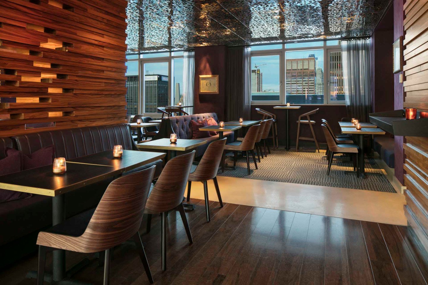 Modern restaurant interior with wooden floors and walls, featuring several tables and chairs arranged neatly. Candles are placed on the tables, adding a warm ambiance. Large windows reveal a cityscape view, with tall buildings visible outside.