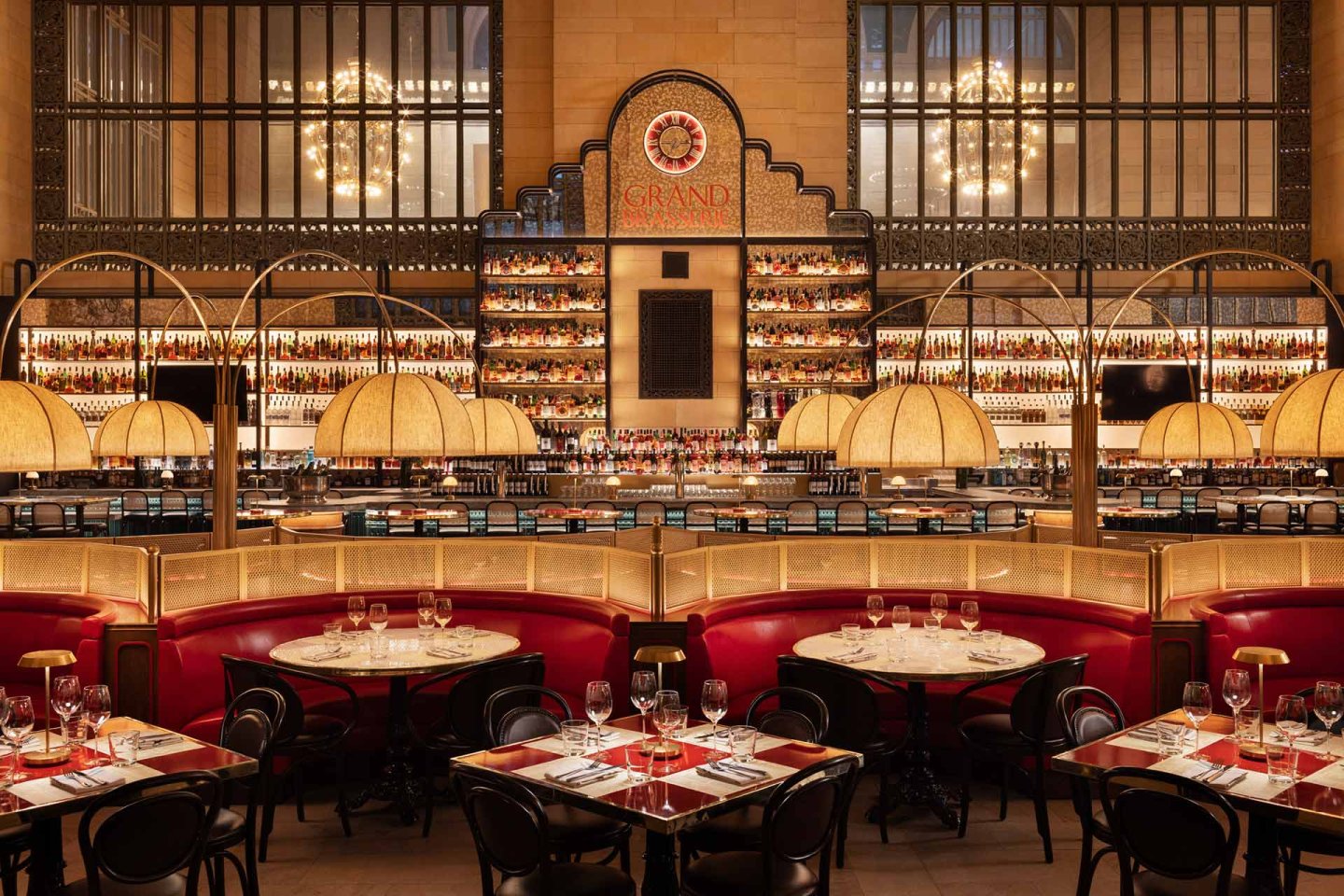 Elegant restaurant interior with red leather booths, round tables set with glasses and cutlery, art deco lighting, and a grand bar with illuminated shelves lined with bottles. Large windows and chandeliers add to the upscale ambiance.