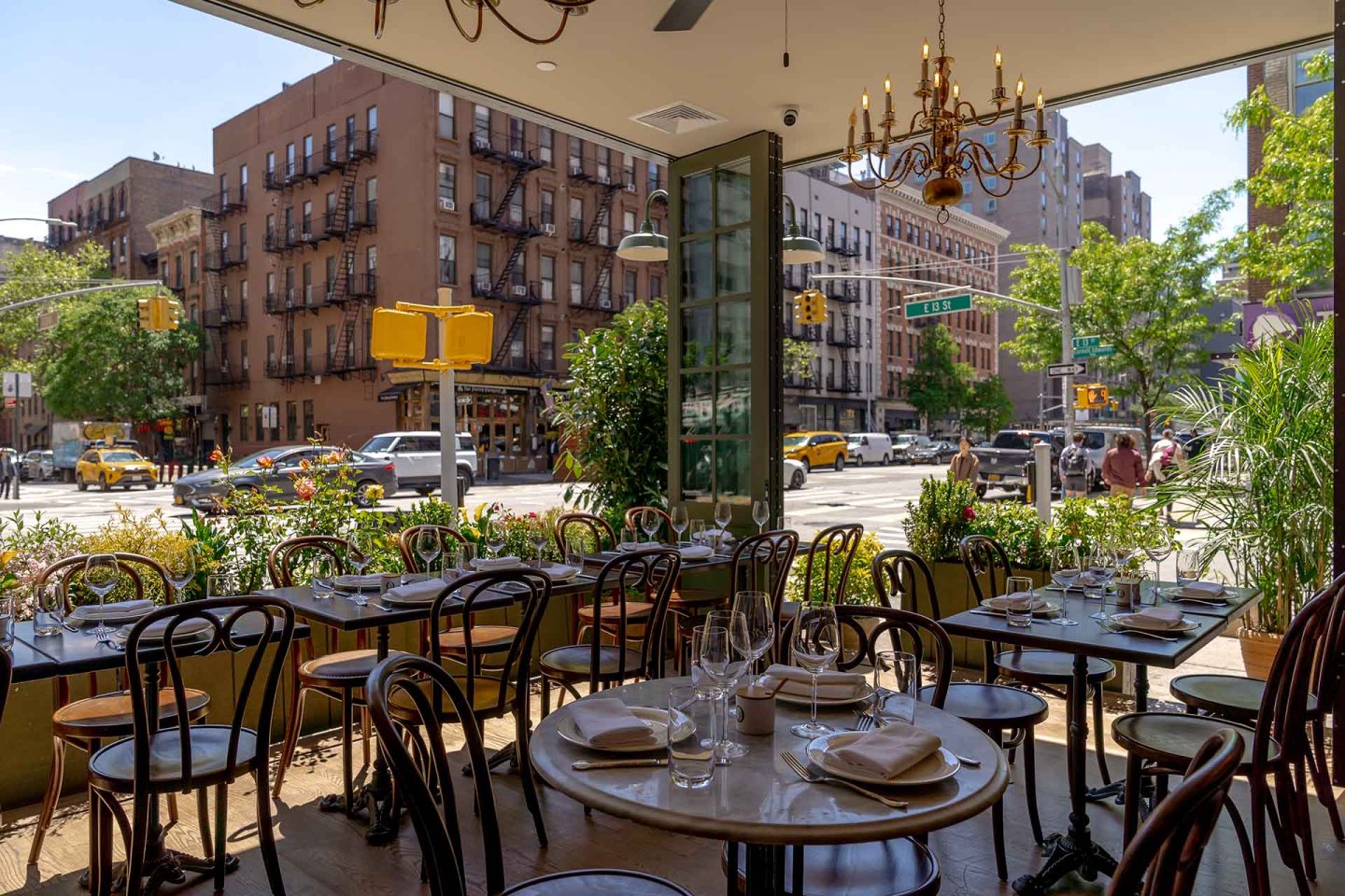 A sunlit restaurant with set tables and wooden chairs faces large open windows, revealing a city street with buildings, greenery, and yellow taxis outside.
