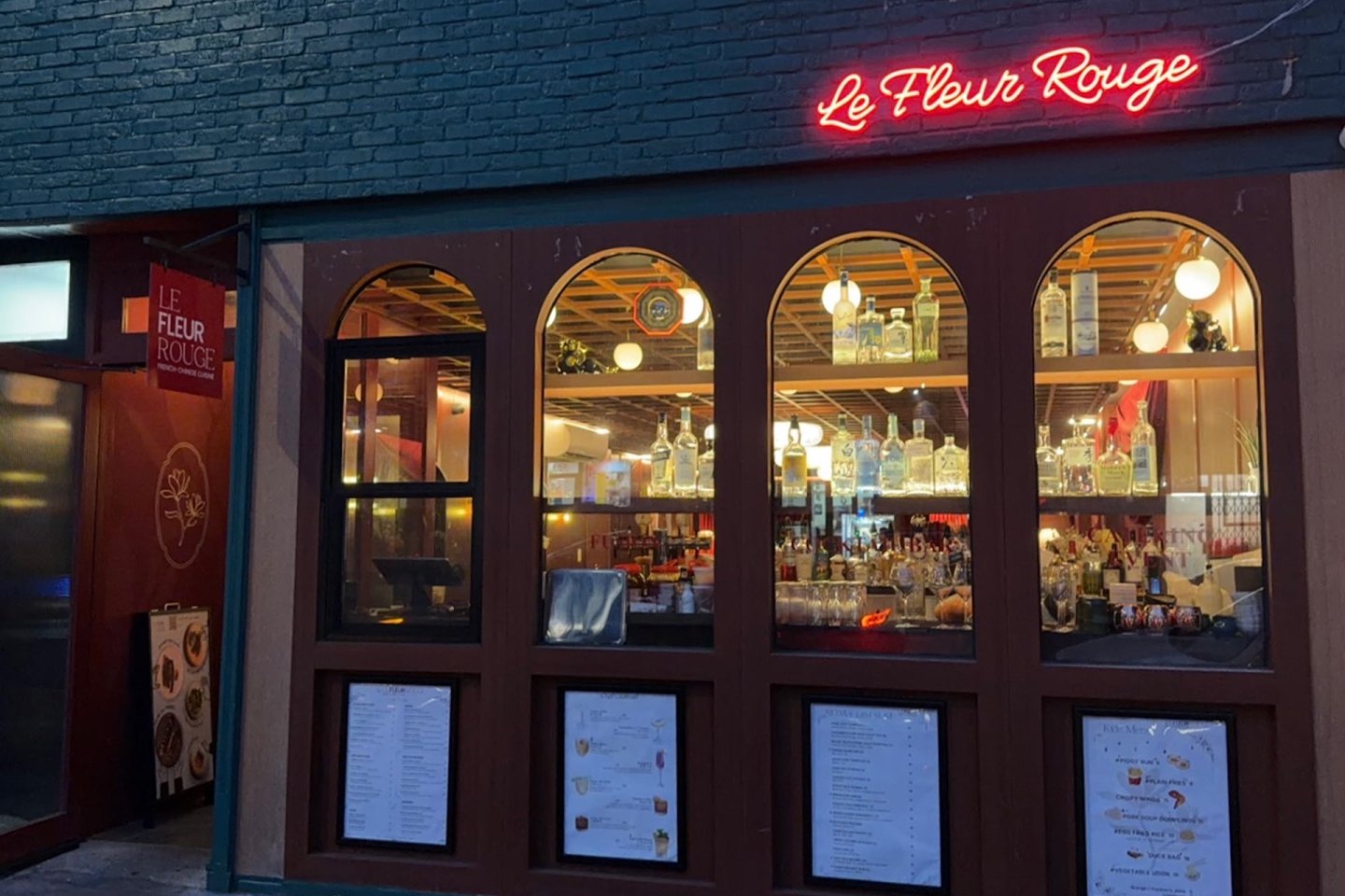 A restaurant called "Le Fleur Rouge" with a neon red sign, large arched windows displaying bottles on shelves, and printed menus posted on the exterior walls. Warm lights are visible inside.