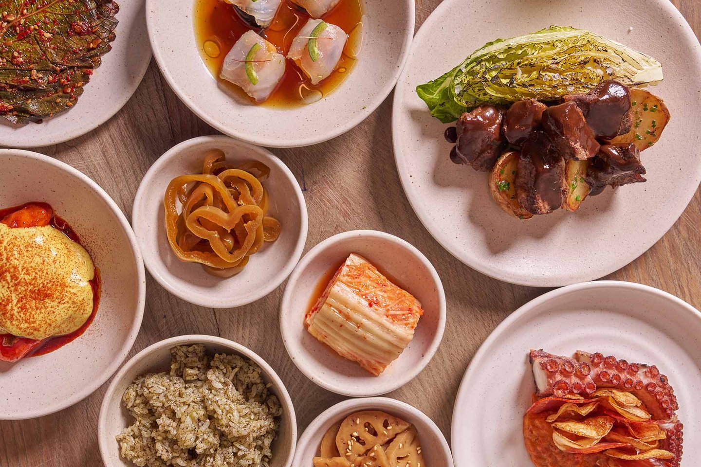 An overhead view of assorted small dishes, including grilled vegetables, sliced meats, seafood, pickles, kimchi, rice, and plates with savory sauces, all arranged on a wooden table.