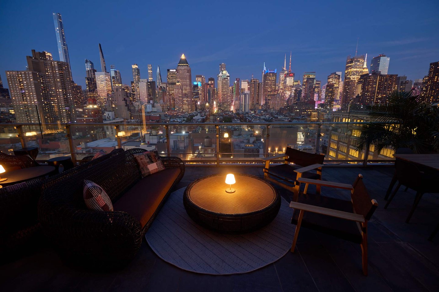 A cozy rooftop lounge area with wicker seating and a round table illuminated by a small lamp, overlooking a brightly lit city skyline at dusk.