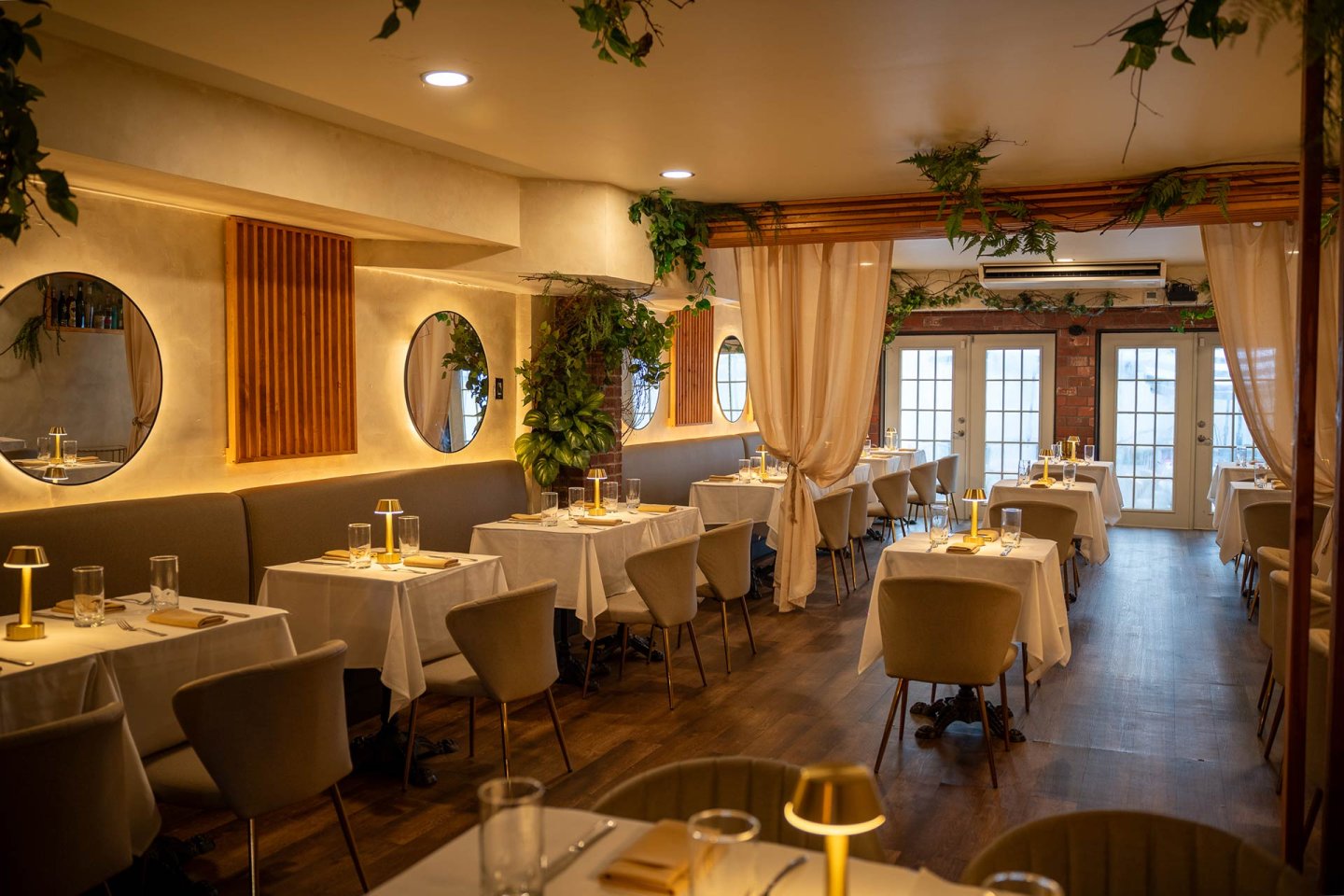 Elegant restaurant interior with round mirrors, white tablecloths, soft beige chairs, warm lighting, greenery, and sheer curtains. Tables are set and arranged in a cozy, inviting atmosphere with wooden floors.
