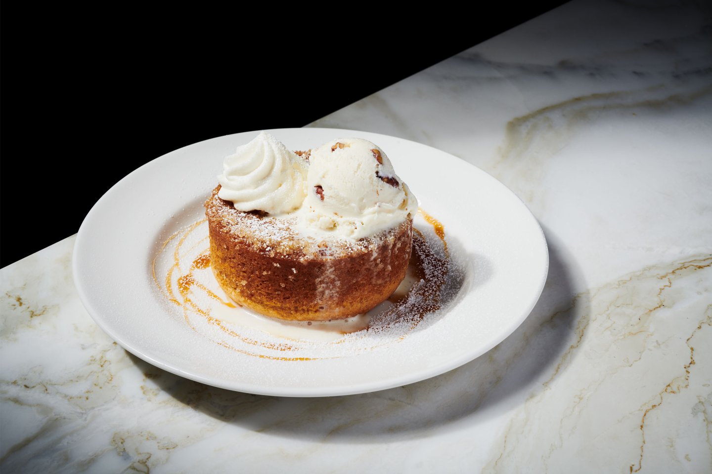 A round dessert topped with whipped cream and a scoop of vanilla ice cream, dusted with powdered sugar, served on a white plate with caramel sauce, placed on a marble surface.