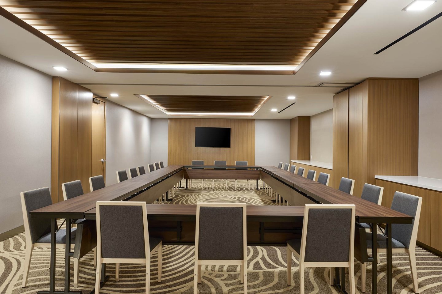 Modern conference room with a U-shaped wooden table, several gray chairs, wood-paneled walls, patterned carpet, a wall-mounted TV, and recessed ceiling lights. The room is empty and well-lit.