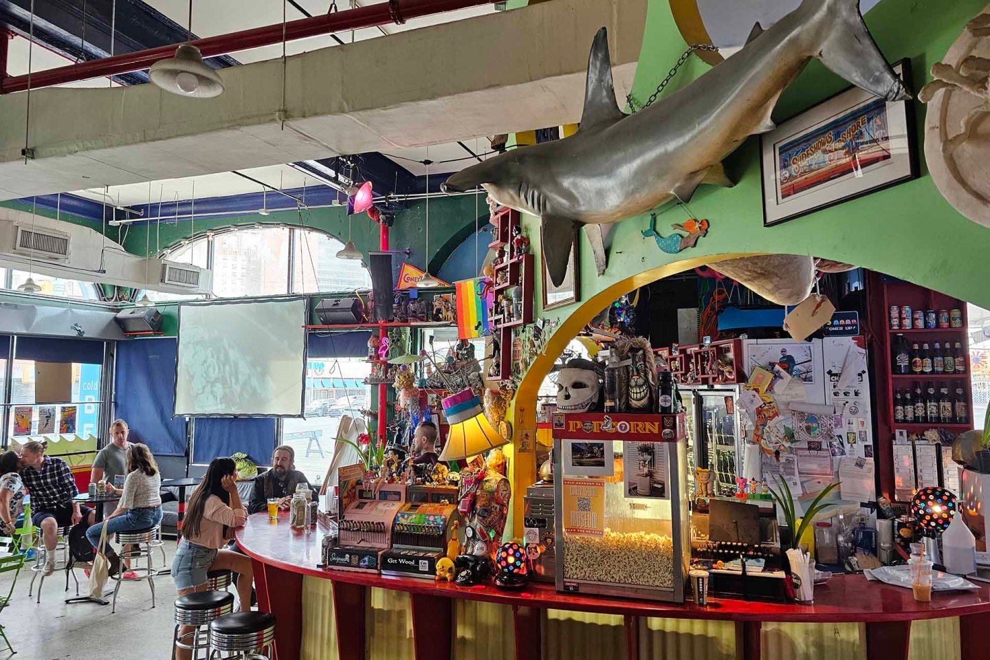 A colorful bar with quirky decor including a large hanging shark, popcorn machine, rainbow flags, and various knick-knacks. Several people sit at tables and the bar, with large windows and a projector screen in the background.