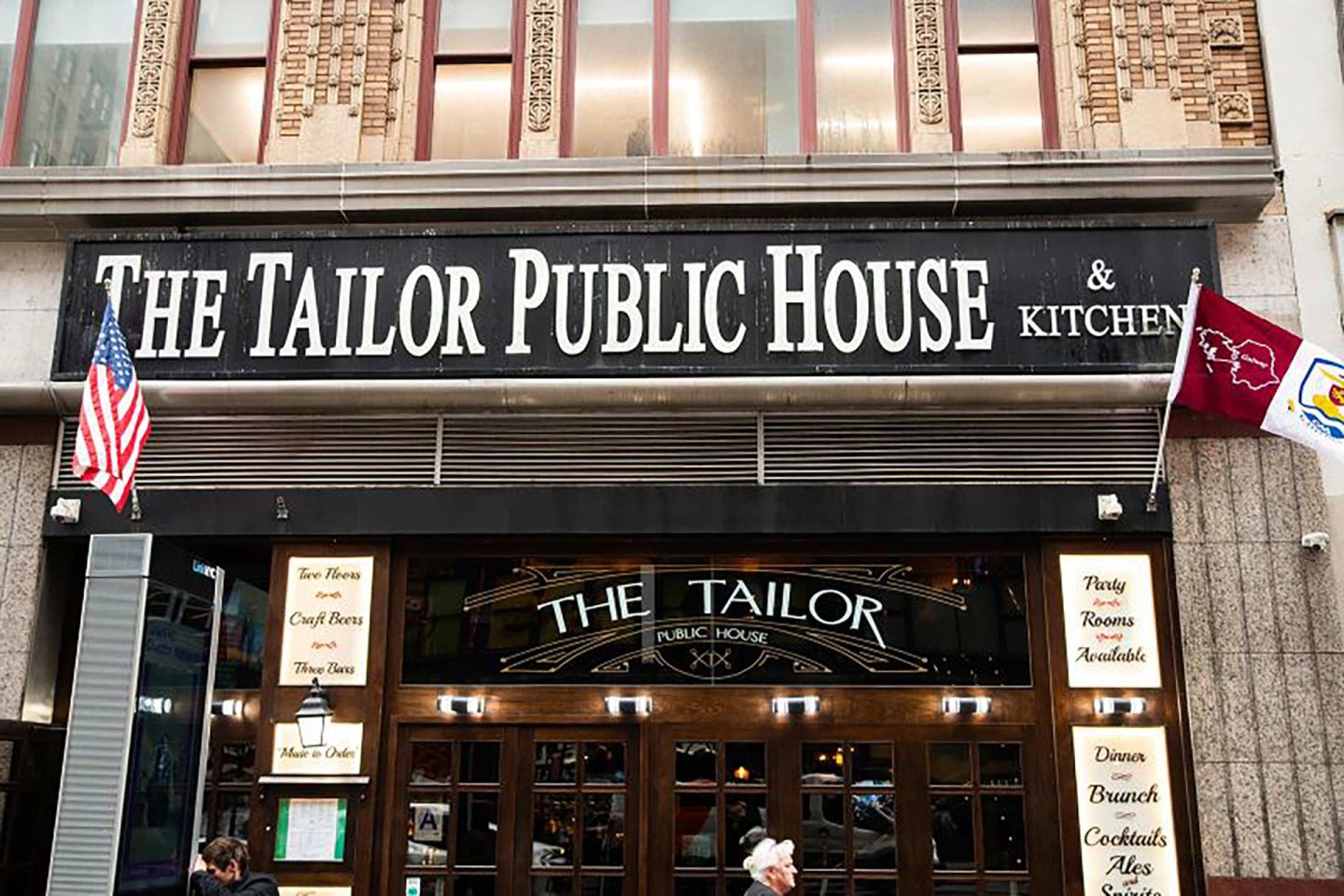 The entrance of The Tailor Public House & Kitchen, featuring a black sign with white lettering, American and maroon flags, and wooden double doors with menus posted outside.