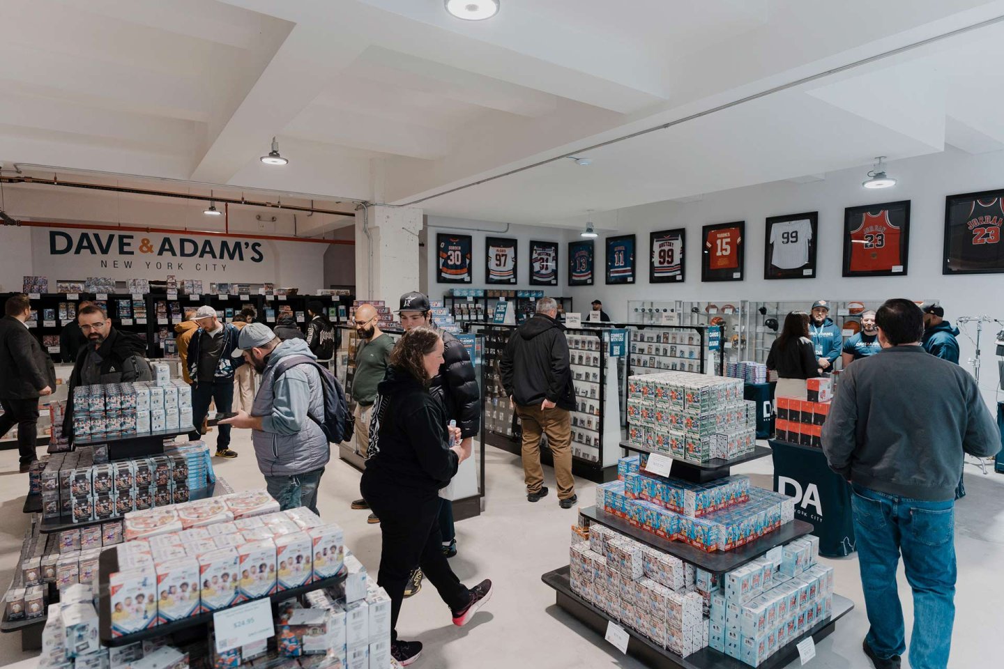 A sports memorabilia store with several people browsing. Shelves are filled with trading card boxes, and framed jerseys hang on the walls. The store sign reads "Dave & Adam's New York City.