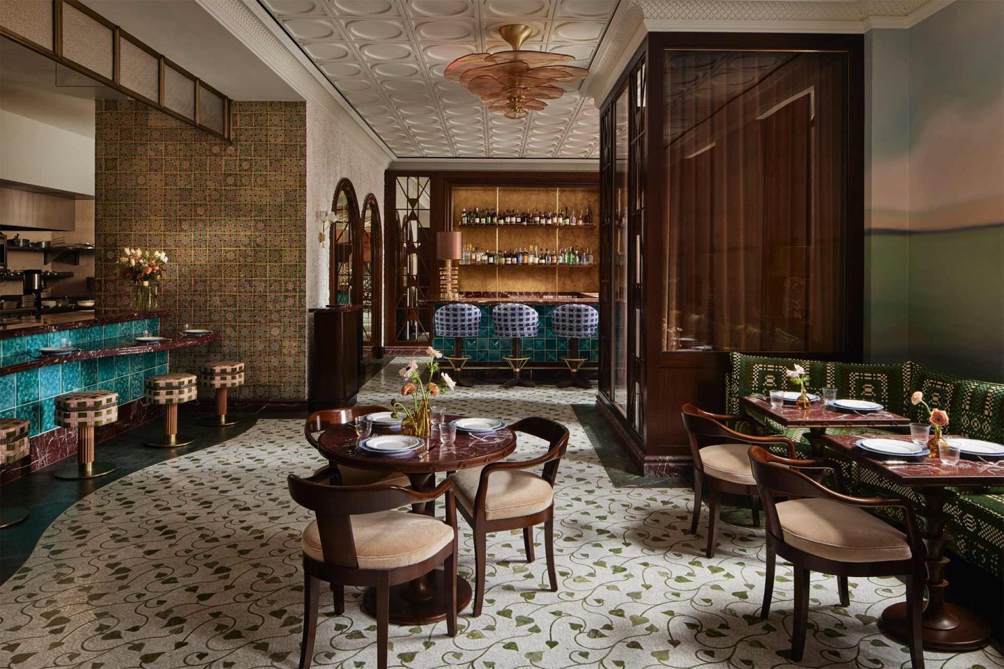 Elegant restaurant interior with patterned floor, wooden tables, cushioned chairs, green banquette seating, a teal-tiled bar, bar stools, and decorative lighting. Shelves with bottles are visible behind the bar area.
